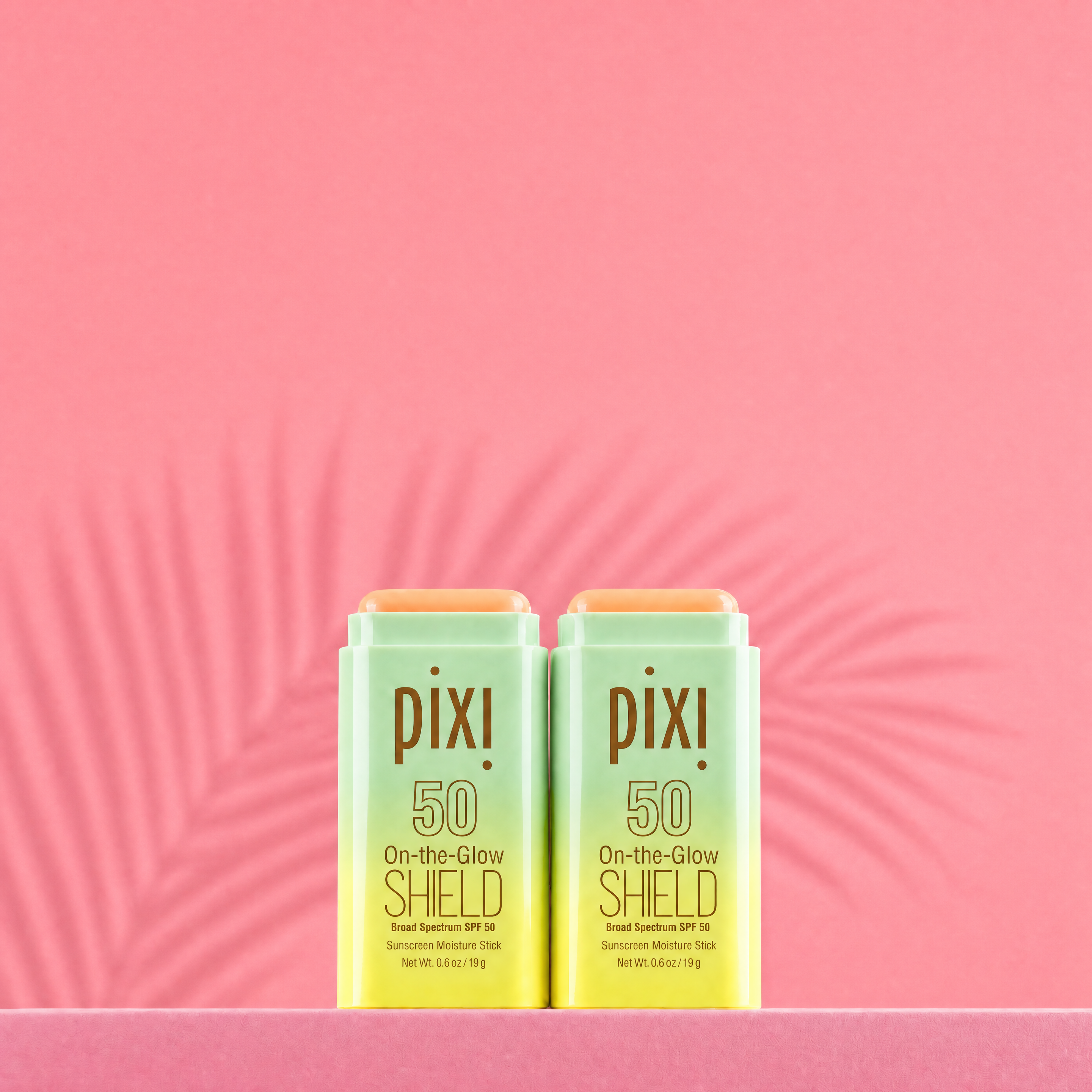 Two Pixi On-the-Glow Shield SPF 50 bottles on a pink background with a shadow of a plant - View 1