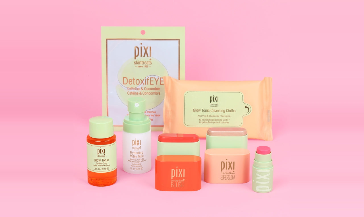 Pixi skincare set arranged on a pink background, including a DetoxiEYE pouch, cleansing wipes, blush, superglow, toner, serum, and a lip treatment. The products feature pastel and bright colors like orange, coral, mint green, and peach.