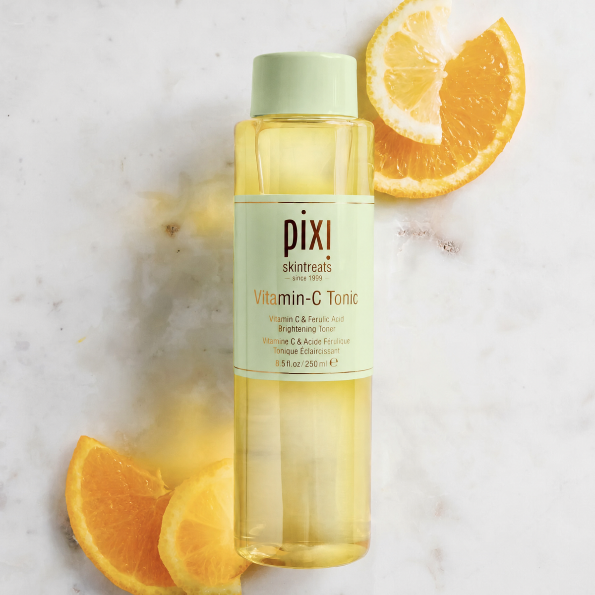 Pixi Skintreats Vitamin-C Tonic in a clear bottle with a pale green cap and label, styled on a white marble surface with orange slices.