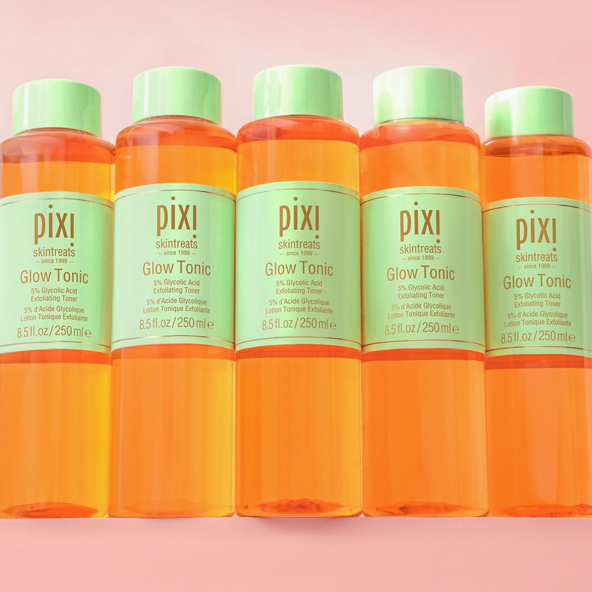 Five bottles of Pixi Glow Tonic arranged in a row against a soft pink background. The bottles are filled with bright orange liquid and have light green caps and labels.