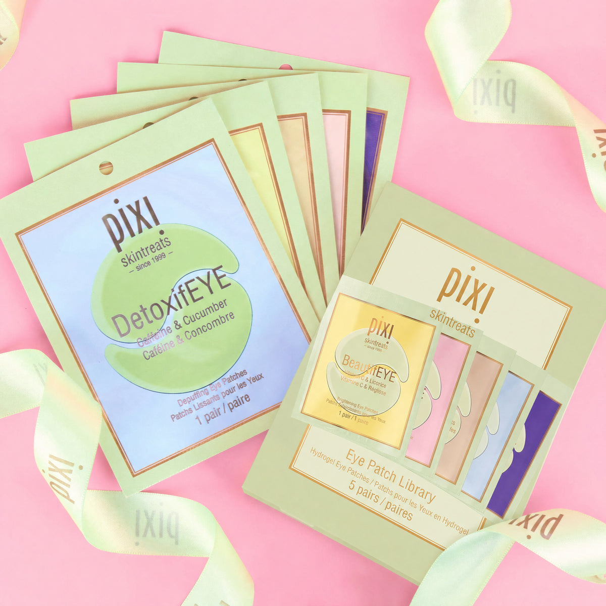 A styled flat-lay of Pixi Skinetreats eye patch sachets on a pink background with pale green ribbon accents. The image shows multiple individual sachet packs fanned out and a boxed 'Eye Patch Library' set, both branded Pixi.