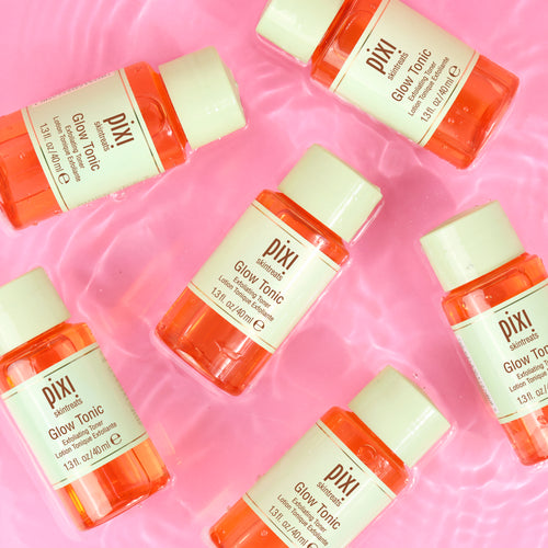Six small Pixi Glow Tonic bottles arranged on a pink water-textured background. The bottles are translucent orange with cream-colored caps and labels, and the brand name Pixi is clearly visible. view 2