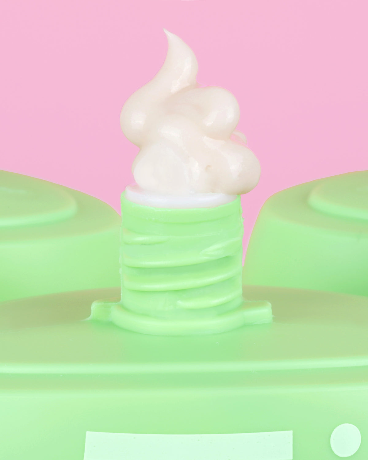 A close-up of Pixi HandHero hand lotion dispensing a swirl of cream against a solid pink background.