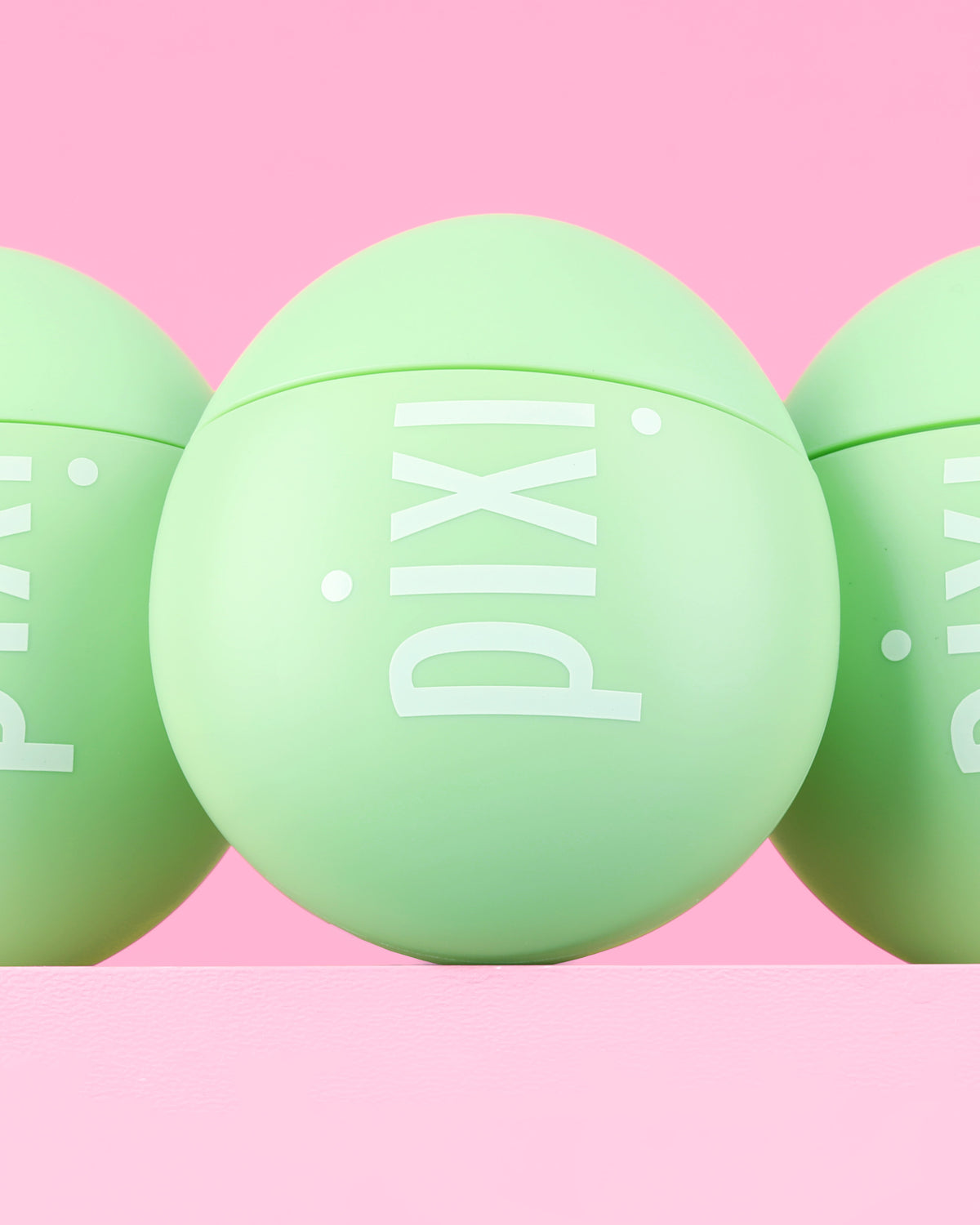 Three round, matte mint-green Pixi HandHero containers arranged in a row against a solid pink background.