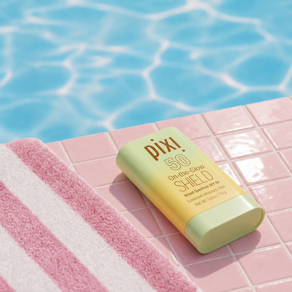 Pixi On-the-Glow Shield SPF 50 sunscreen sits on a pink tiled poolside surface next to a pink and white striped towel, with blue pool water in the background.