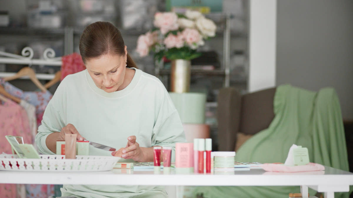 A woman in a light sage green sweatshirt sits at a white table, applying makeup from a small pink tube to her hand. Several cosmetic products in pink, red, and white packaging are arranged on the table in a softly lit room with a blurred background of clothing racks and flowers.
