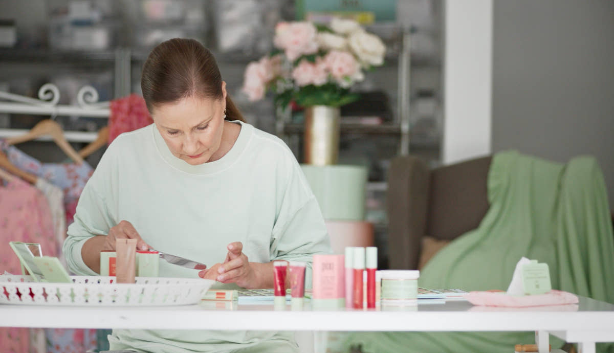 A woman in a light sage green sweatshirt sits at a white table, applying makeup from a small pink tube to her hand. Several cosmetic products in pink, red, and white packaging are arranged on the table in a softly lit room with a blurred background of clothing racks and flowers.