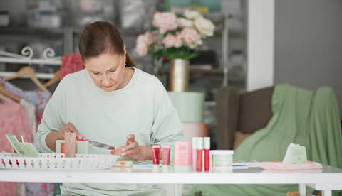 A woman in a light sage green sweatshirt sits at a white table, applying makeup from a small pink tube to her hand. Several cosmetic products in pink, red, and white packaging are arranged on the table in a softly lit room with a blurred background of clothing racks and flowers.