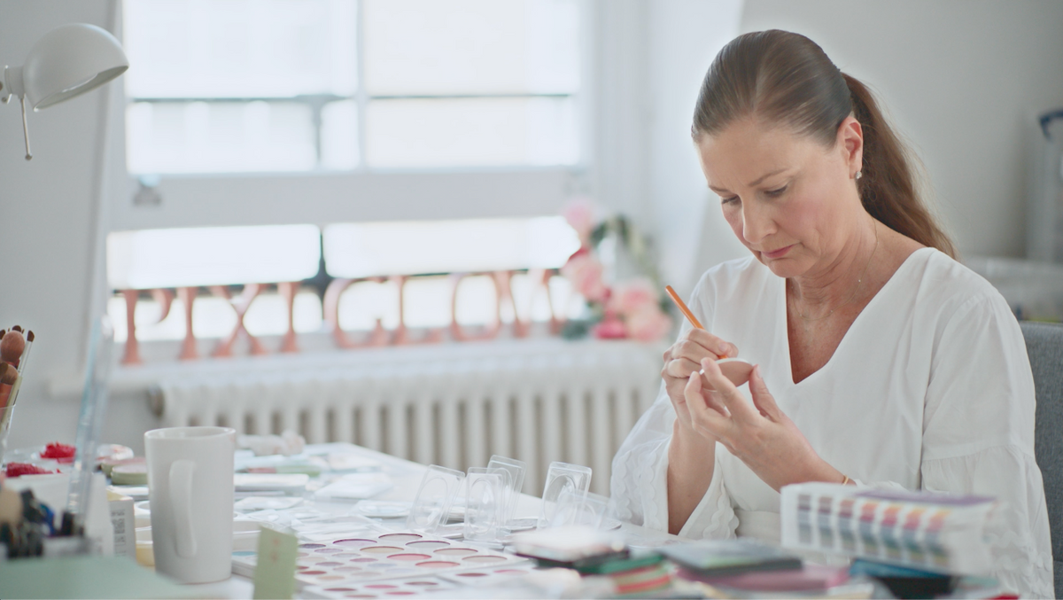 Petra Strand, Pixi Founder, with her hair in a ponytail sits at a table, carefully examining a small clear object with a pencil. The room is bright and airy with a large window in the background, and the table is cluttered with various craft or sample materials.