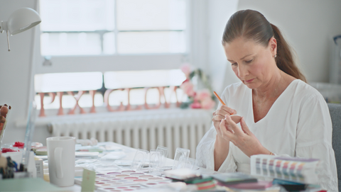 Petra Strand, Pixi Founder, with her hair in a ponytail sits at a table, carefully examining a small clear object with a pencil. The room is bright and airy with a large window in the background, and the table is cluttered with various craft or sample materials.