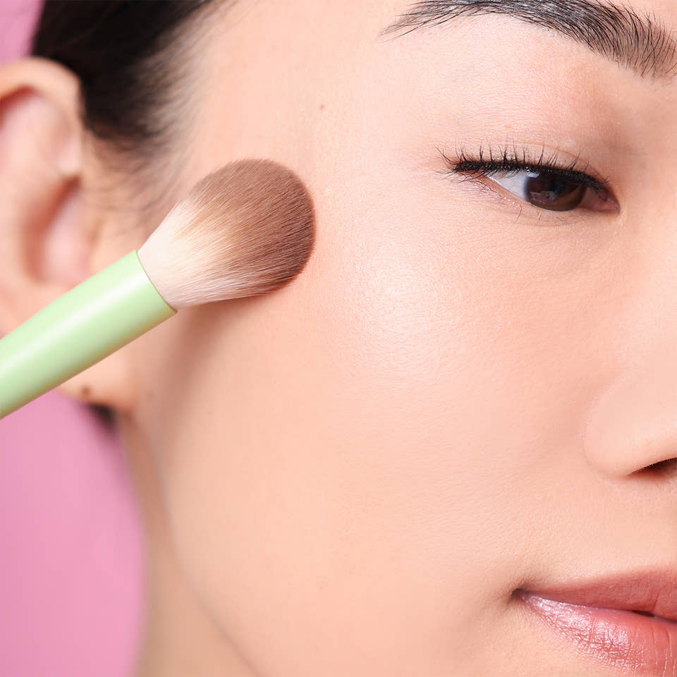 Close-up of a person applying makeup with Pixi Glow Brush on a blurred pink background view 1