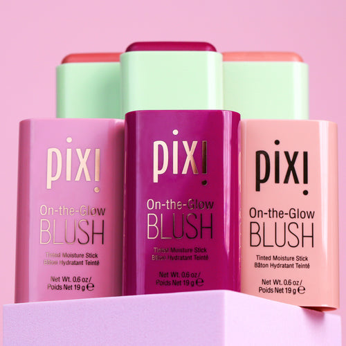 Three Pixi On-the-Glow Blush tinted moisture sticks in pink, purple, and peach packaging, arranged on a pastel pink background with a lavender pedestal. view 1