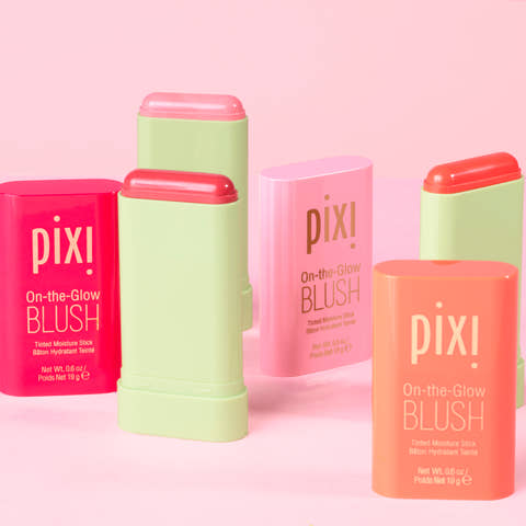 Pixi On-the-Glow Blush sticks in assorted pastel and bright shades, arranged in a clean studio setup against a solid pink background.