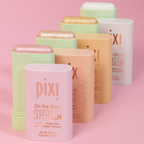 Five Pixi On-the-Glow SuperGlow hydrating moisturizer sticks in assorted pastel shades, arranged in a neat diagonal row on a solid pink background.