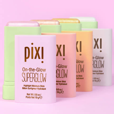 Four Pixi On-the-Glow SuperGlow highlight and moisture sticks in pastel shades of pink, peach, orange, and mint green, arranged in a row against a solid pink background.