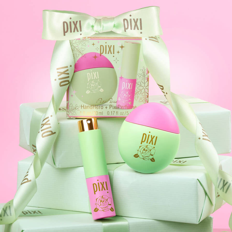PixiRose Perfume and HandHero gift set arranged on stacked light green boxes with a pale green ribbon bow. The set includes a round mint-green and pink rollerball, a matching tube with a pink lower section and gold cap, and a printed gift box insert, all against a soft pink background. view 1