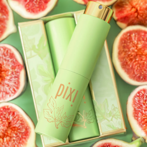 A Pixi cosmetic spray bottle in light green with a gold cap, displayed diagonally on top of its matching green box, surrounded by sliced pink figs on a green background. view 2
