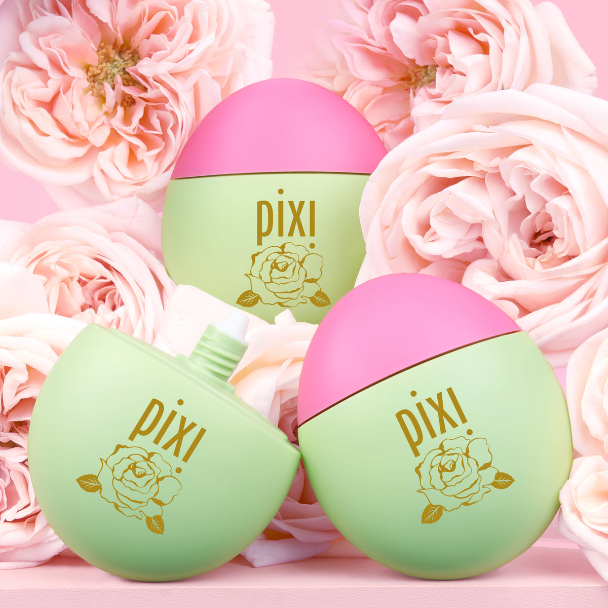 Three Pixi HandHero PixiRose scented products arranged among pink roses on a pink background. One cap is removed to show applicator tip.