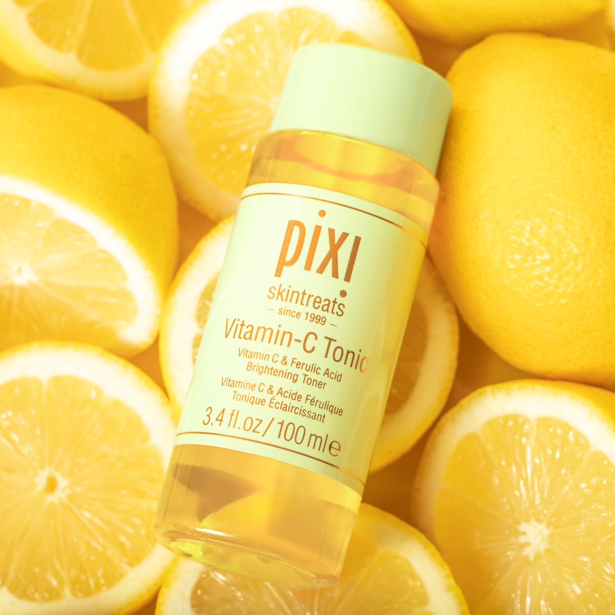 Pixi Skintreats Vitamin-C Tonic bottle with a light green cap, surrounded by fresh yellow lemon slices and whole lemons.