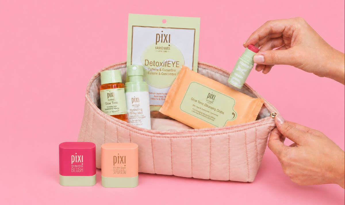 A pink quilted cosmetic pouch filled with Pixi skincare products, including a DetoxiEYE packet, Glow Tonic Petite, Hydrating Milky Mist Petite, Glow Tonic Cleansing Cloths, Hydra LipTreat, On-the-Glow BLUSH and SUPERGLOW Petites in front on a solid pink background. A hand is reaching in to adjust the pouch zipper.