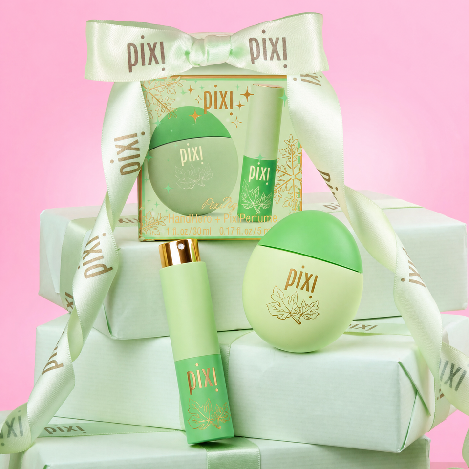 PixiPerfume gift set featuring a round lip balm container, a cylindrical tube with a gold cap, and a decorative box wrapped with a white satin ribbon, all in light green and gold packaging against a pink background. view 1